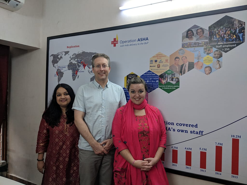 Tableau volunteers Michael and Lauren with Operation ASHA Director Sonali in front of a poster in the Operation ASHA offices